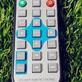MP5 Remote control MP4 Video Player Remote Price - a rectangular grey remote with a blue navigation cross, volume buttons, and playback controls on grass.