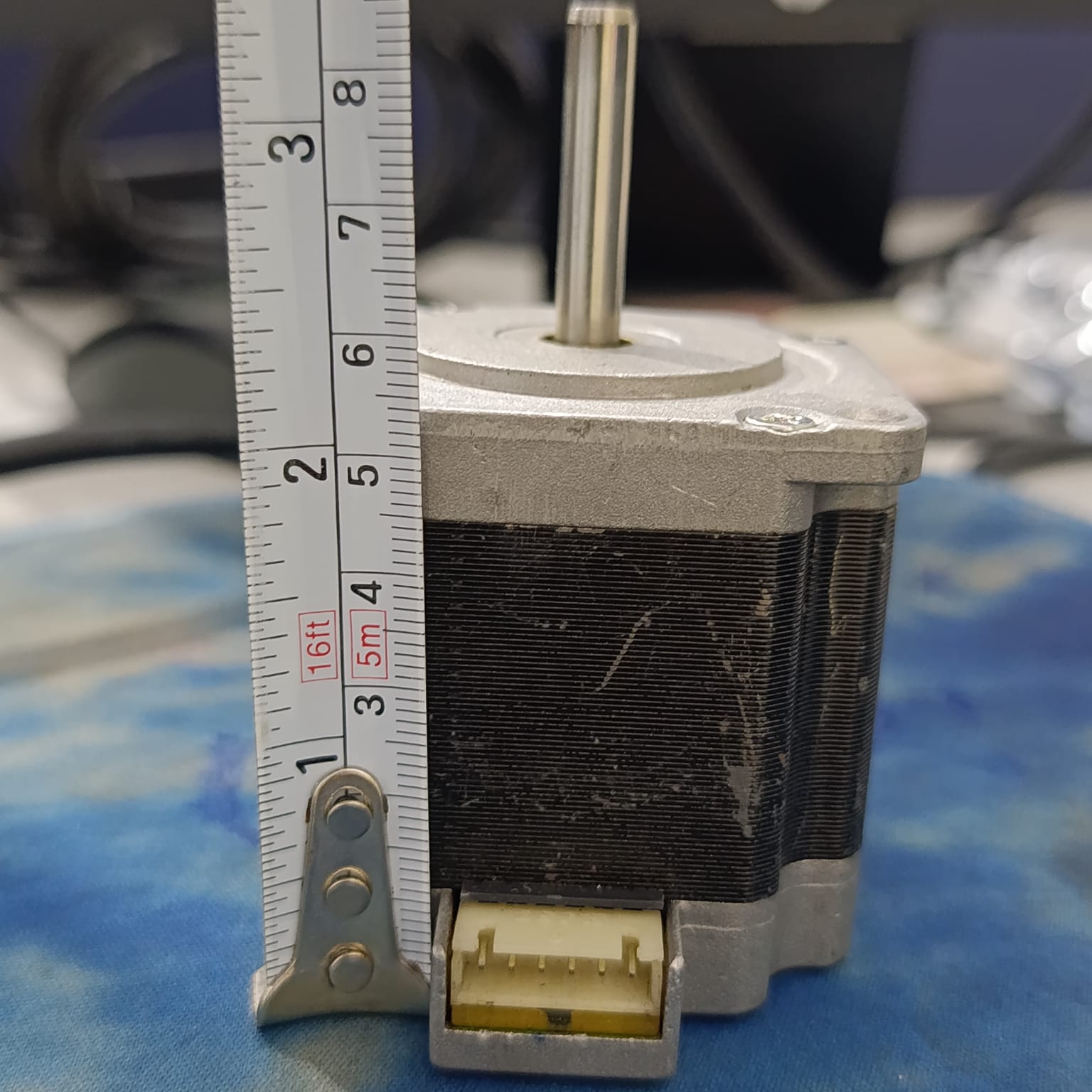 NEMA 23 Stepper Motor In Pakistan (1.5A–2.0A, 1.8°) - a silver and black square motor with a top shaft and a bottom white connector, shown next to a metal measuring tape.