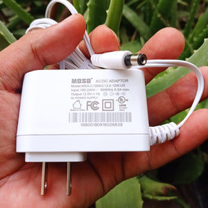 New High Quality 220V To 12V 1A AC DC Adapter - a white wall-plug power supply with a barrel connector held in a hand against a green plant background.