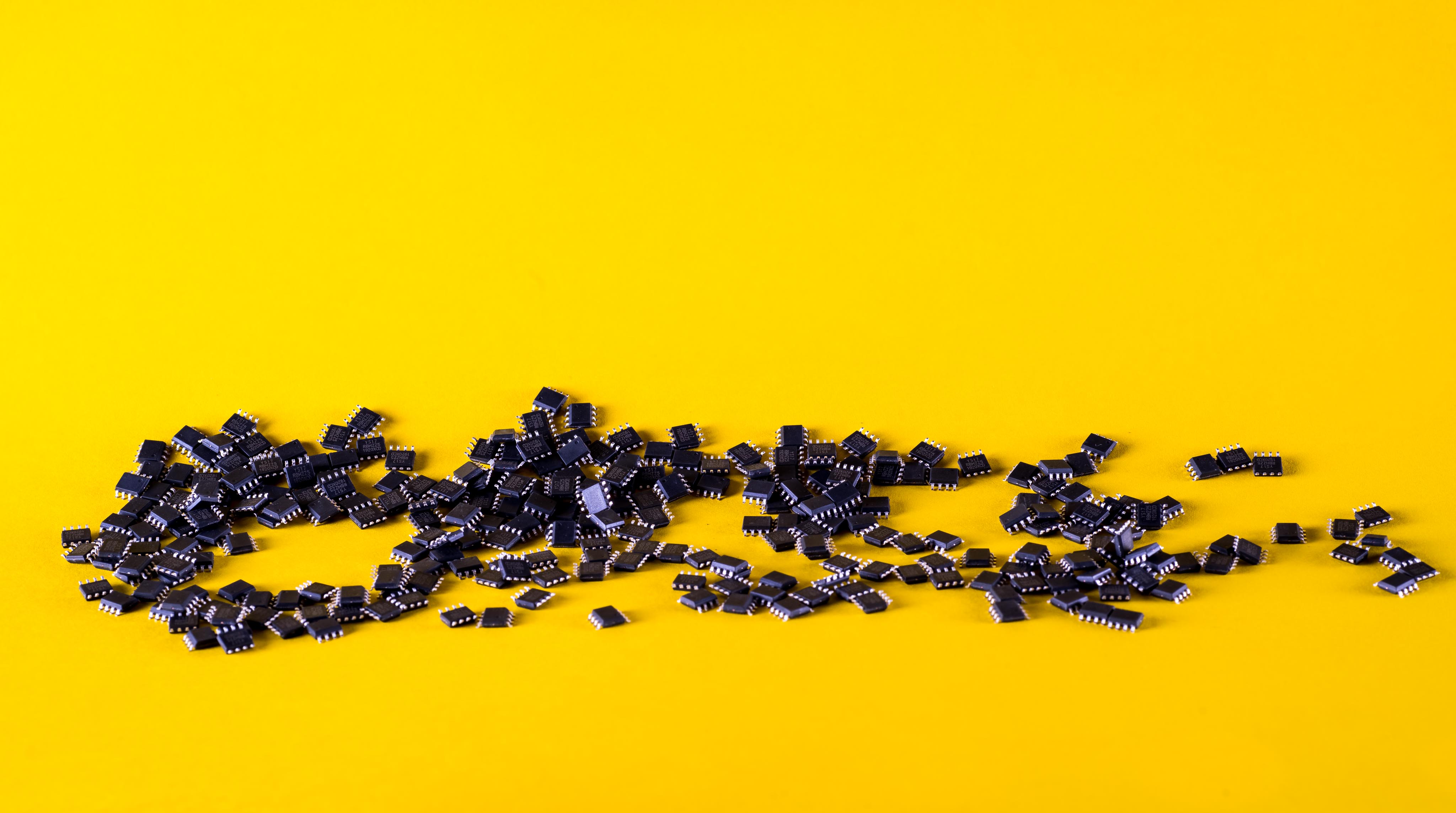On Demand Cheapest Microcontroller - a large pile of small black integrated circuit chips with metallic pins scattered across a bright yellow background.
