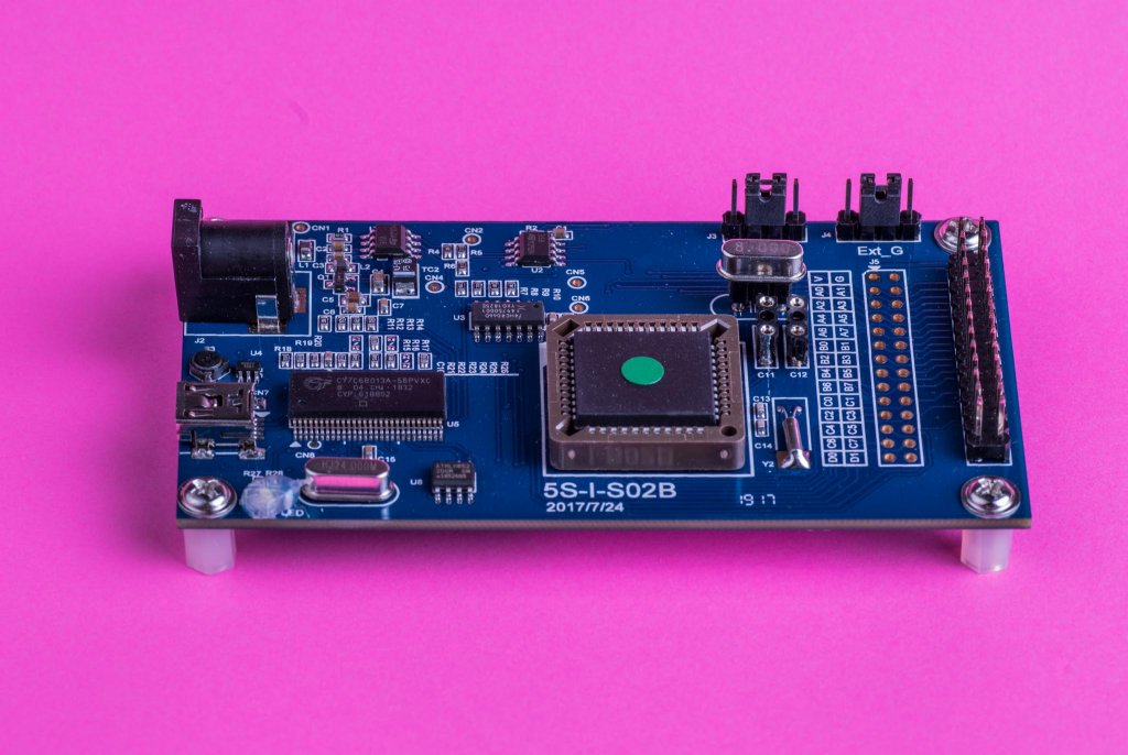 On Demand Cheapest Microcontroller - a blue rectangular circuit board featuring a central chip, a power jack, and multiple pin headers on a pink surface.