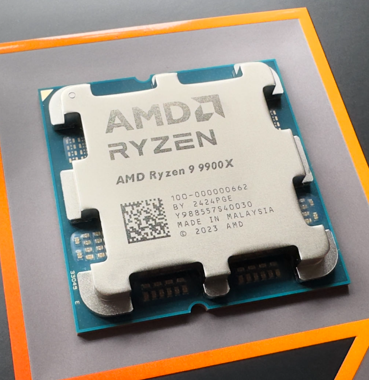 On Demand R9 9900X Processor 12-Core, 24-Thread - AMD Ryzen 9 9900X processor, a silver-coloured square CPU with an integrated heat spreader, resting on a flat surface with orange-bordered packaging.