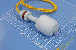 P45 Vertical Mount Float Switch For Water Level Sensing - a white plastic liquid level sensor with a red O-ring and yellow wires, resting on a centimeter measurement grid.