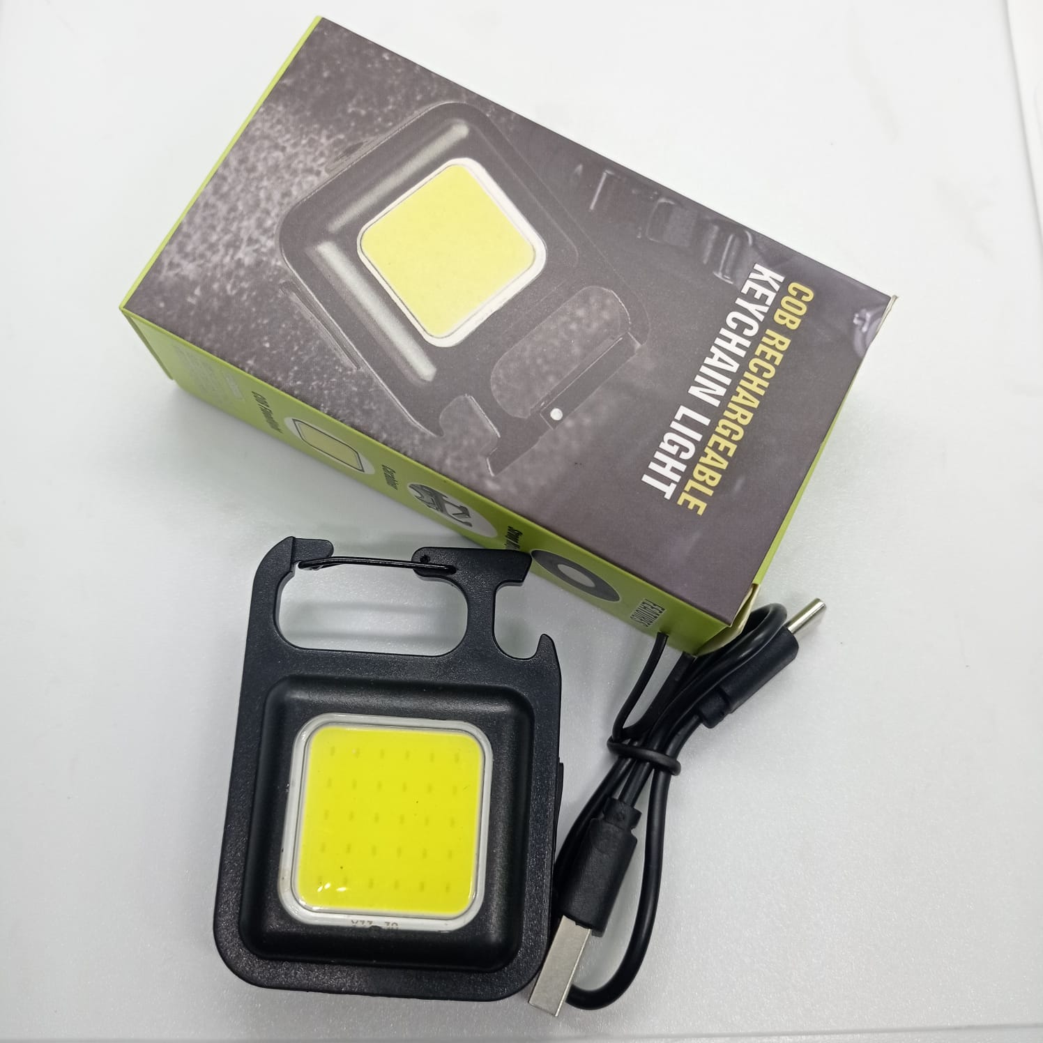 Portable COB Flashlight - Emergency Light with Rechargeable Keychain - Durable Plastic Casing - with its black box packaging, a USB charging cable, and a black square light unit featuring a carabiner clip and bottle opener.
