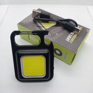 Portable COB Flashlight - Emergency Light with Rechargeable Keychain - Durable Plastic Casing - black square casing, integrated carabiner, and a USB charging cable resting on its product packaging box.