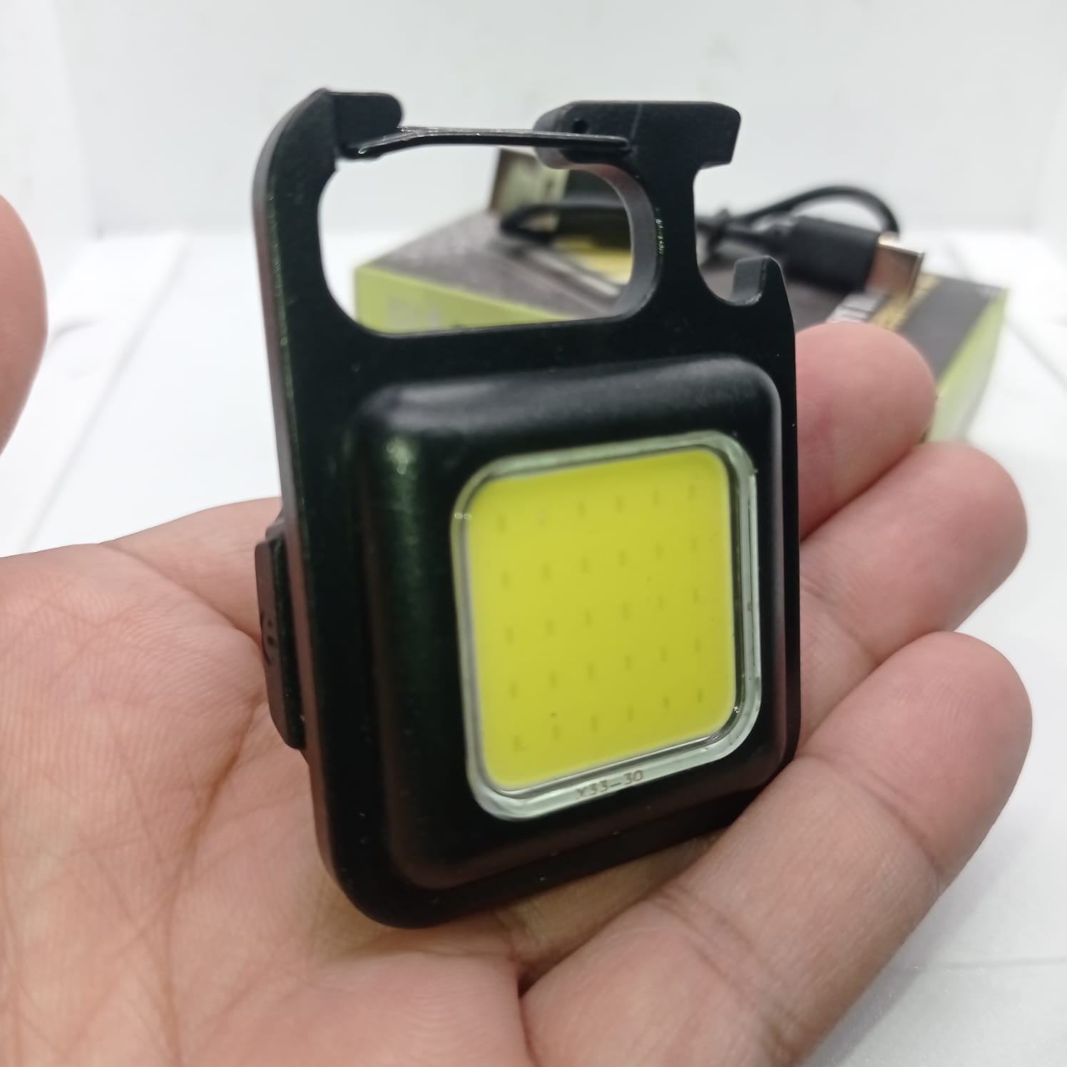 Portable COB Flashlight - Emergency Light with Rechargeable Keychain - Durable Plastic Casing - held in a hand, featuring a black square casing with a yellow light panel, a carabiner top, and a charging cable in the background.