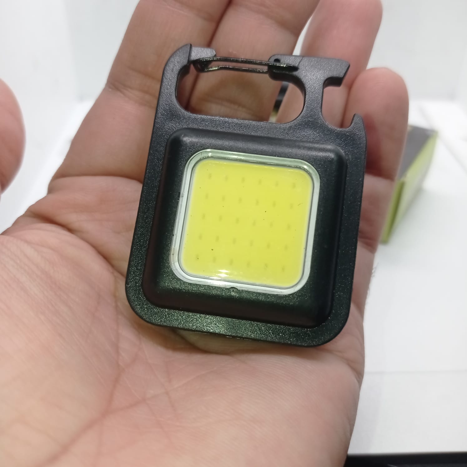 Portable COB Flashlight - Emergency Light with Rechargeable Keychain - Durable Plastic Casing - a black square-shaped rechargeable keychain light with a COB LED panel and integrated carabiner held in a hand.