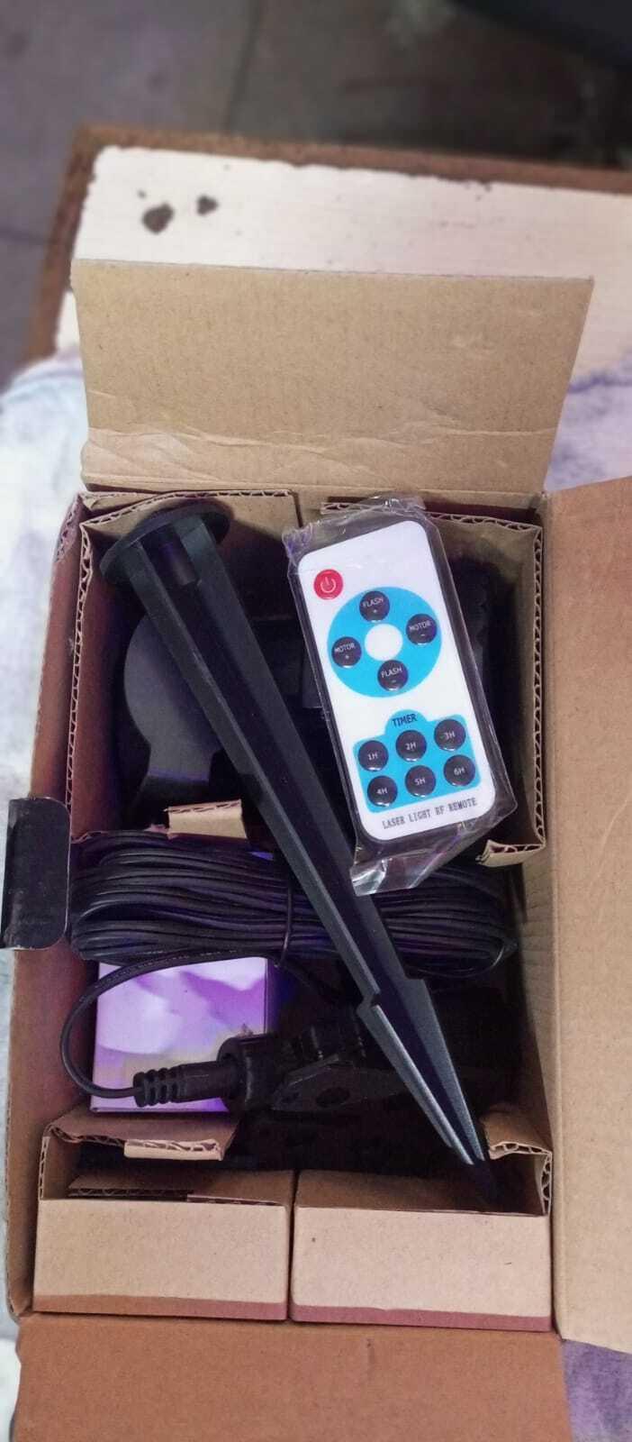 Projector - shown in its cardboard packaging with a black plastic ground stake, a power cord, and a white remote control with timer and flash buttons.
