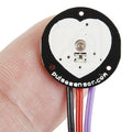 Pulse Sensor Pulse Heart Rate Sensor Arduino Heartbeat Sensor - a close-up of the circular heart-shaped black and white PCB held by fingers with three attached red, black, and purple wires.