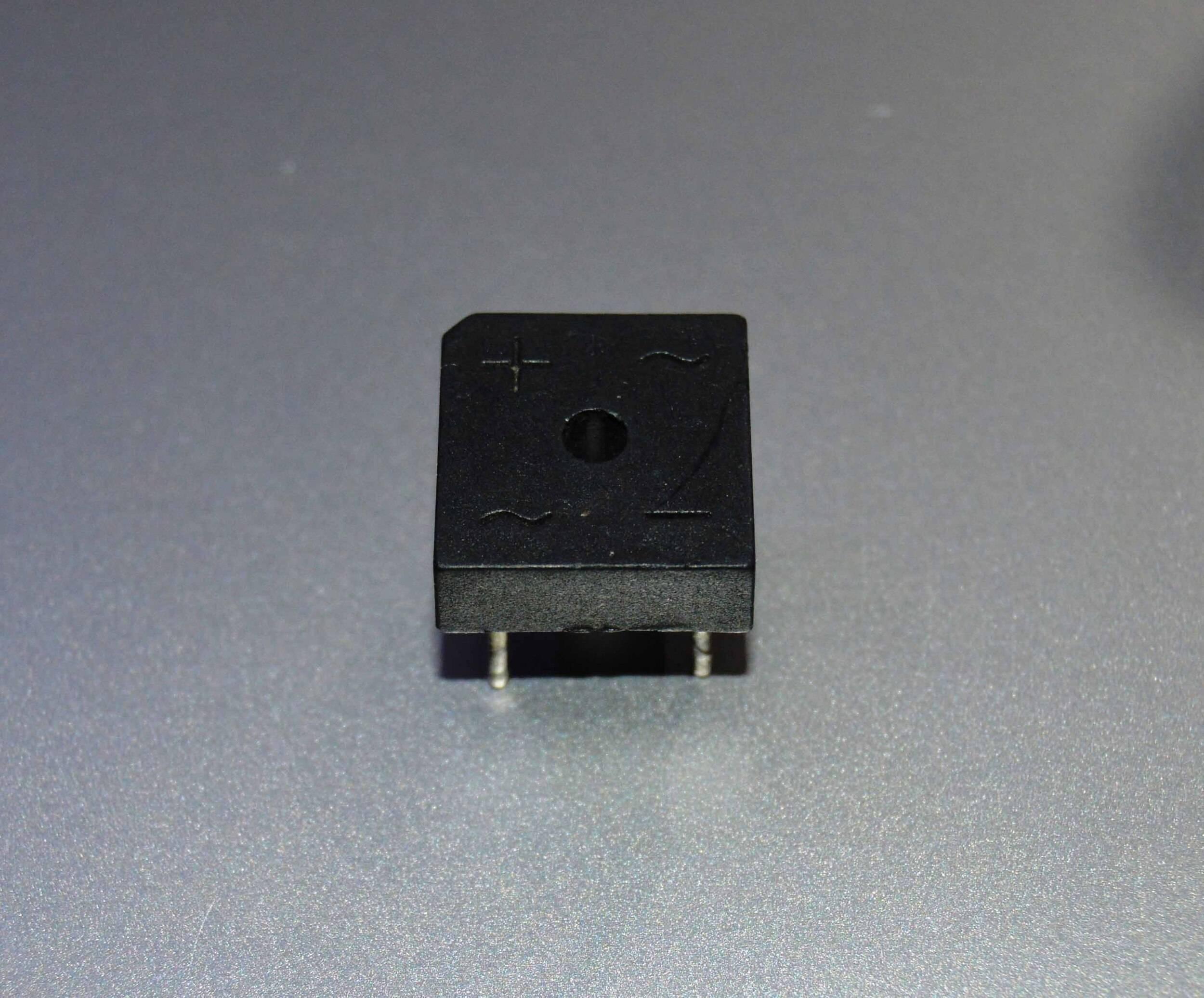 Rectron Bridge Ractifier BR104 (used Condition) - a black, square-shaped electronic component with four metal pins and symbols for positive and AC inputs on its surface.