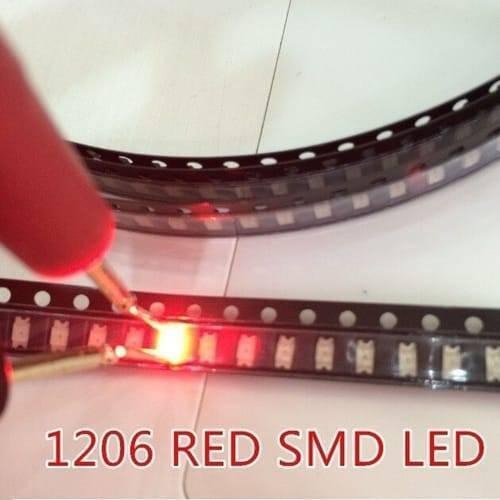 Red SMD 1206 LED Super Bright Light Emitting Diode - a glowing red surface-mount light-emitting diode being tested with probes while housed in a strip of black plastic carrier tape.