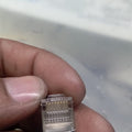 RJ45 Connector with meteal casing - with metal casing, shown in a close-up view held by fingers, displaying the transparent plastic housing and the eight internal gold pins.