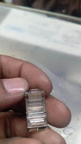 RJ45 Connector with meteal casing - with metal casing, shown in a close-up view held by fingers, displaying the transparent plastic housing and the eight internal gold pins.
