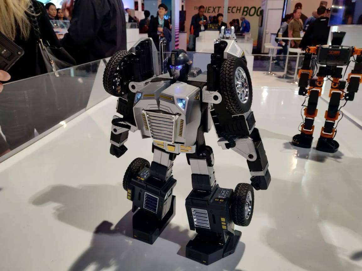 Robosen T9 - Advanced Programmable And Convertible Robot - STEM: Fun, Educational With Voice And App Control - a silver and black humanoid robot with wheel-shaped shoulders standing on a white surface with another robotic figure in the background.