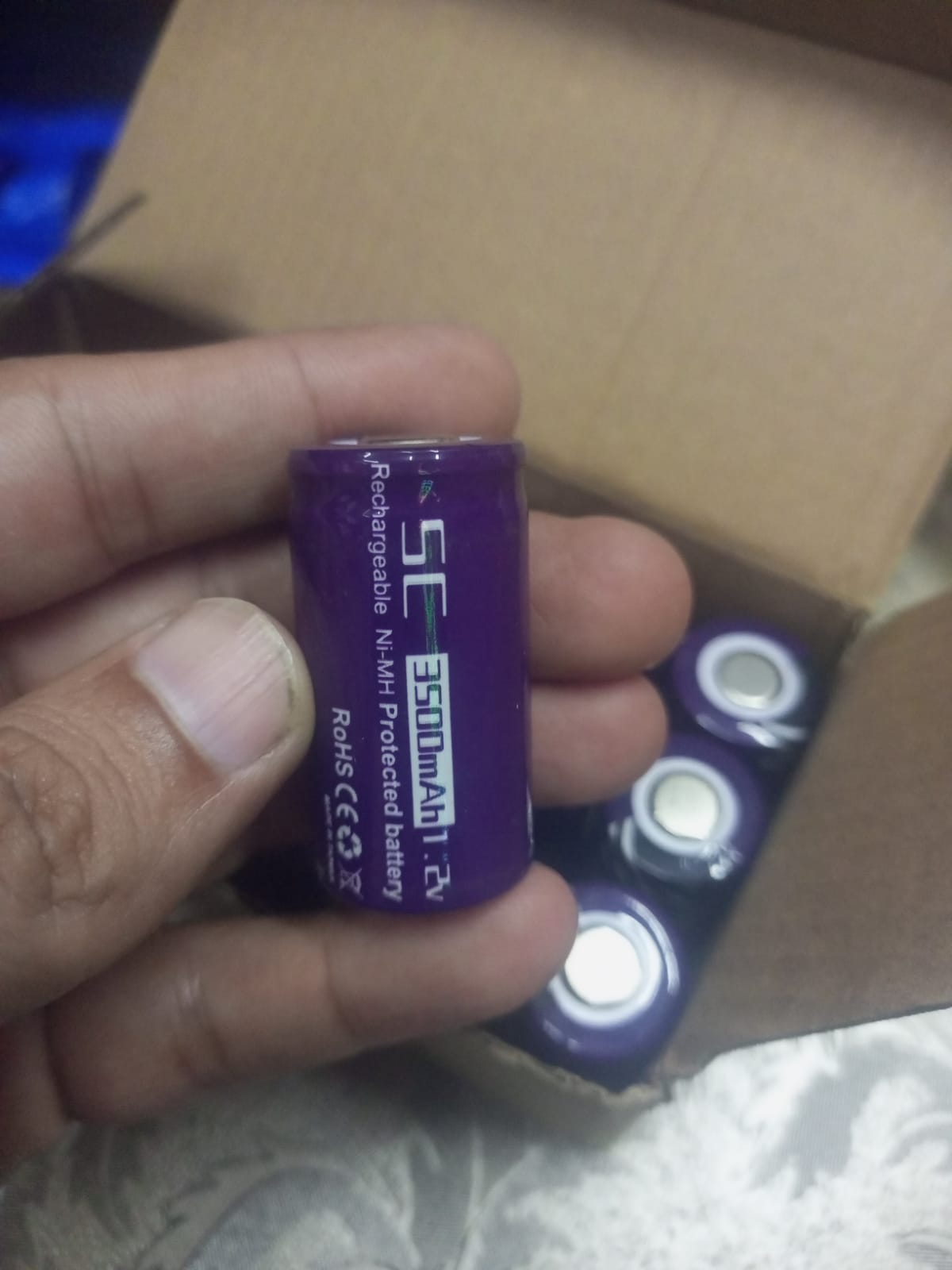 SC 3500mAh 1.2V NiMH Rechargeable Battery - a purple cylindrical cell held in a hand with a cardboard box containing additional batteries in the background.