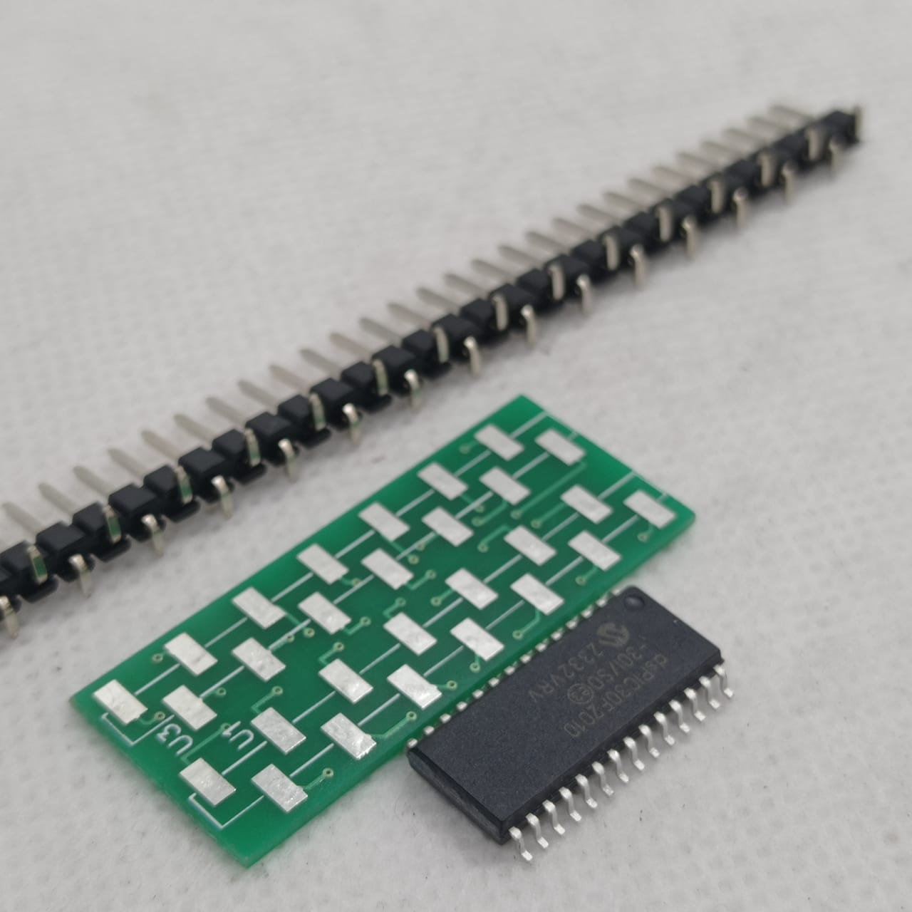 SMD DSPIC30F2010 - microcontroller chip, a green breakout adapter board, and a long strip of header pins arranged on a white surface.