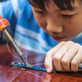 Soldering Iron 30WATT - shown in use with a red handle and metal tip, being held by a person soldering a small blue electronic circuit board.