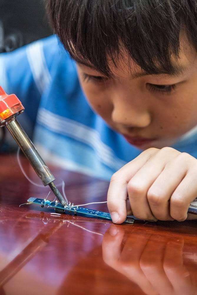 Soldering Iron 30WATT - shown in use with a red handle and metal tip, being held by a person soldering a small blue electronic circuit board.