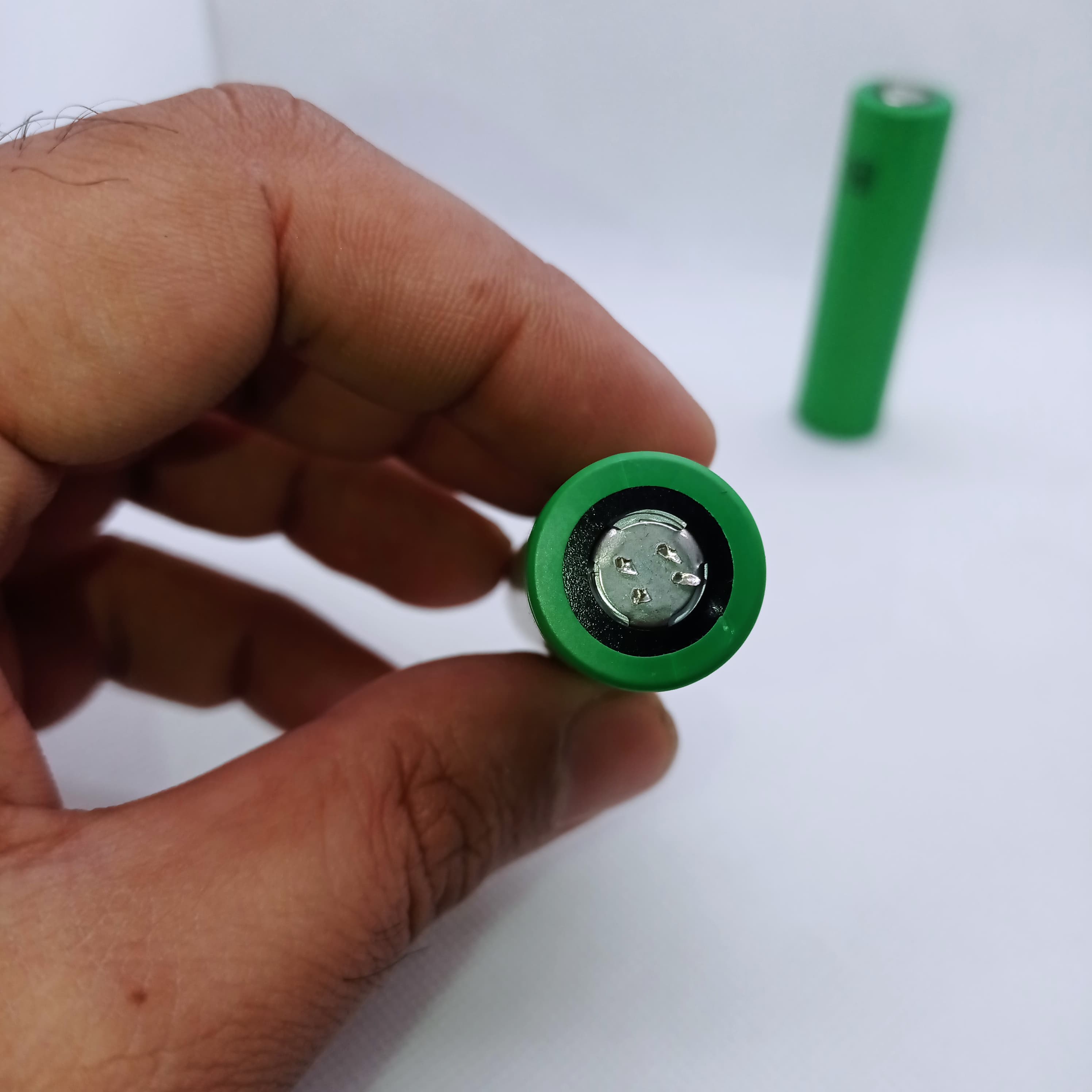 Stock Lot US18650UTC4 3.7V 2000mA 18650 Li-ion Rechargeable Battery – High-Capacity Power Cell for Flashlights, Laptops, and Power Tool - a close-up of the green cylindrical cell's positive terminal held in a hand with another in the back.