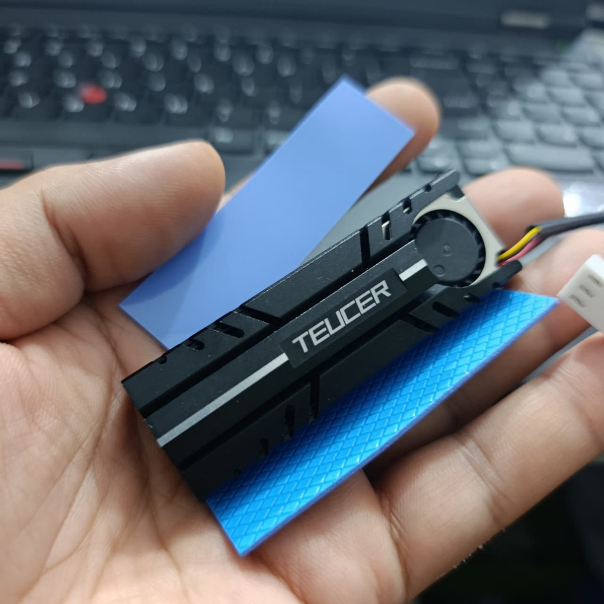 TEUCER M.2 SSD Heatsink NVME 2280 - Aluminum Cooling Radiator for Desktop PC & PS5 - a black aluminum cooler with a small fan, a power cable, and two blue thermal pads held in a hand.