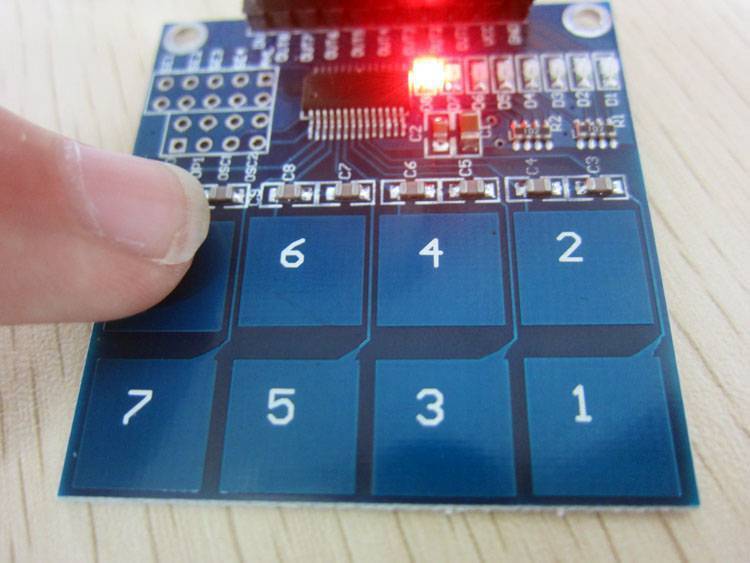TTP226 8 Channel Digital Capacitive Touch Sensor Module - a blue PCB with eight numbered touch pads and a red LED illuminated while being pressed.