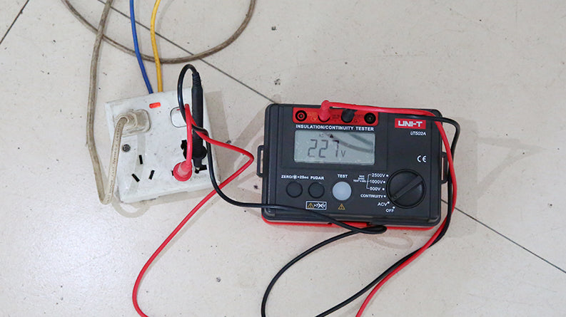 UNI-T UT502A Insulation Resistance Tester – Digital Megohmmeter, 500V/1000V/2500V - connected to a wall socket with red and black leads, showing a 227V reading on its digital display.