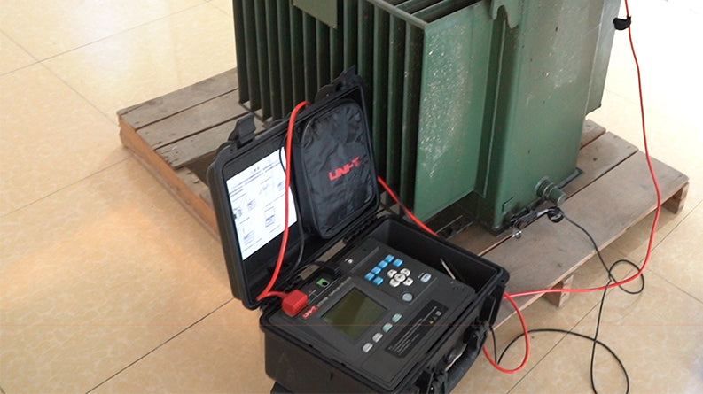 UNI-T UT516B Insulation Resistance Tester – Digital Megohmmeter - black carrying case with a display, keypad, and red cables connected to a green transformer on a pallet.