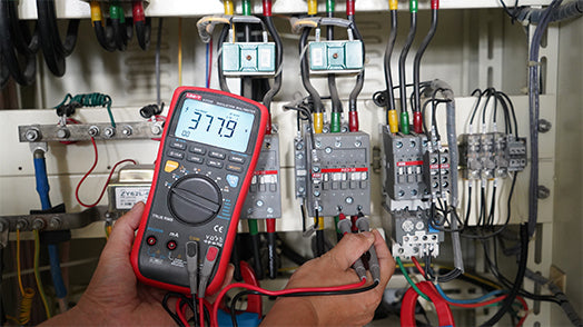 UNI-T UT533 Insulation Multimeter – Insulation Tester + Digital Multimeter - a red handheld device with a digital screen displaying 377.9, held by hands testing electrical components in a panel.