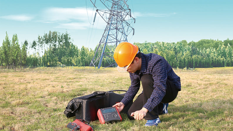 UNI-T UT572 Earth Resistance Tester – Digital Earth / Ground Resistance Meter - a red and black handheld device with a central display and rotary knob, sits on grass next to a black carrying bag.