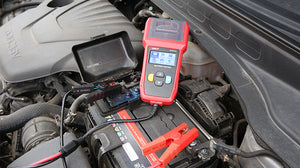 UNI-T UT675A Automotive Battery Tester – 12V/24V, CCA 40–2000, Cranking & Charging System Test, Internal Resistance, Battery Health Analyzer - with a screen showing a menu, connected by cables with red and black clamps to a car battery under the hood.