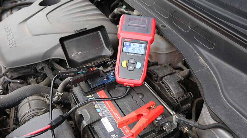 UNI-T UT675A Automotive Battery Tester – 12V/24V, CCA 40–2000, Cranking & Charging System Test, Internal Resistance, Battery Health Analyzer - with a screen showing a menu, connected by cables with red and black clamps to a car battery under the hood.