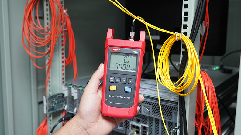 UNI-T UT692G High-Power Optical Power Meter – -50~+26DBM, 800–1700nm, FC/SC/ST, InGaAs, ±5%, IP65, Fiber Optic Power Meter - UNI-T UT692D optical power meter held in a hand, showing a red handheld device with a digital screen reading -70.00 dBm and connected to yellow fiber.