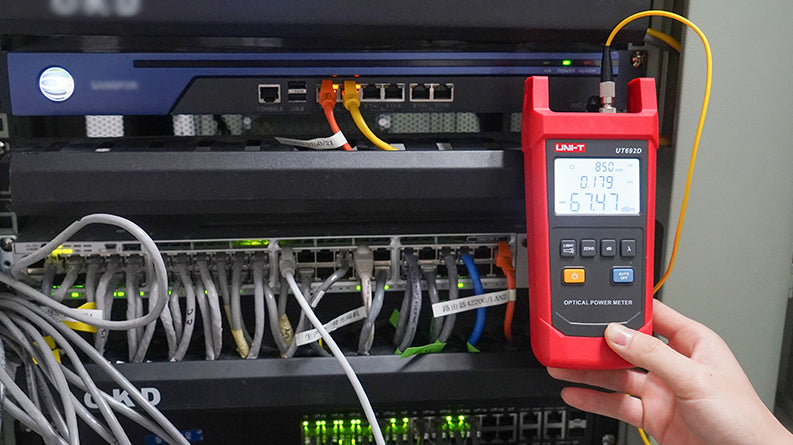 UNI-T UT692G High-Power Optical Power Meter – -50~+26DBM, 800–1700nm, FC/SC/ST, InGaAs, ±5%, IP65, Fiber Optic Power Meter - a red handheld device with a screen showing 850nm and -67.47 dBm, connected by a yellow fiber cable in a server rack.