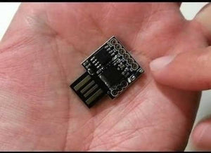 USB Digispark ATTINY85 Developing Board - shown as a small black circuit board with a gold-plated USB connector, held in a person's palm.