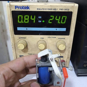 Used 1 Piece DC Solenoid Electromagnet In Mix Shape - held in hand before a Protek PWS-3003D power supply showing 0.84A and 24.0V on its digital display.
