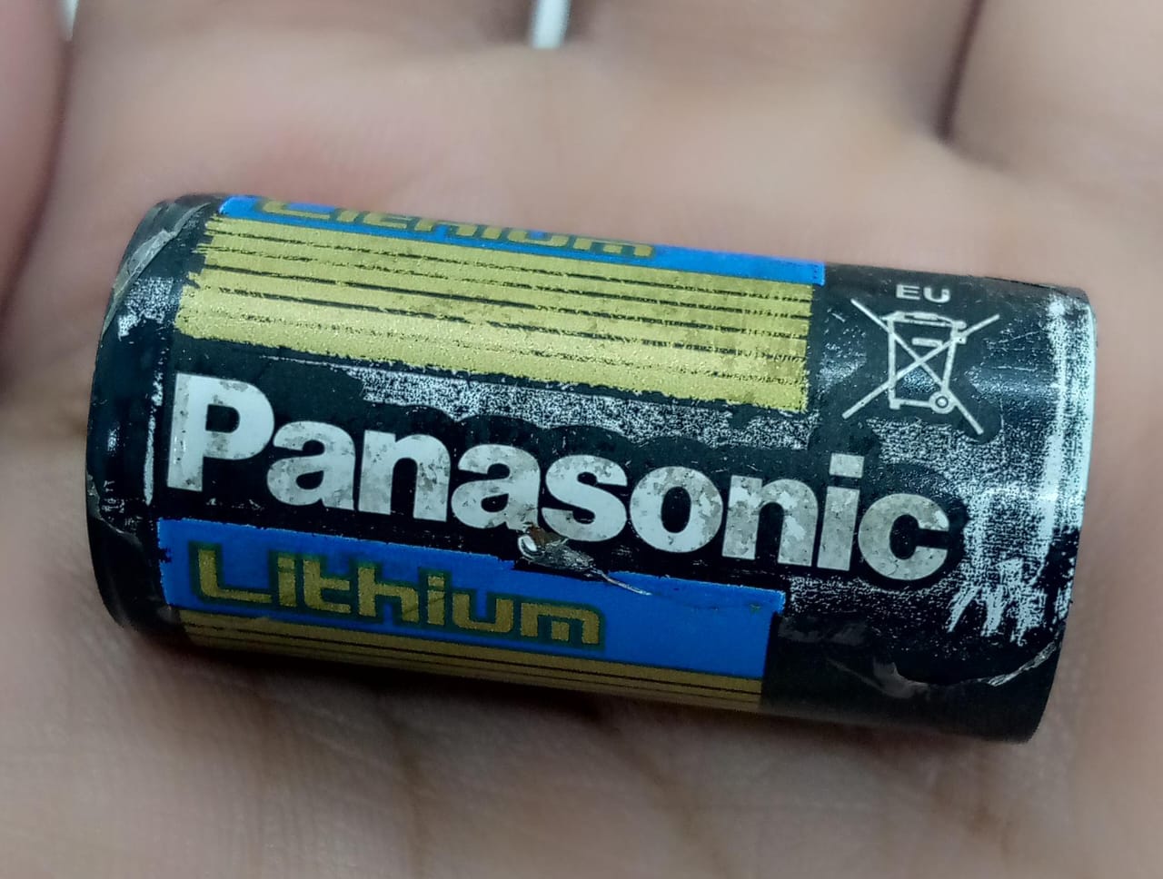 USED Product Panasonic Industrial CR123A Lithium Battery - a black and blue cylindrical cell held in a hand with visible wear and an EU recycling symbol.