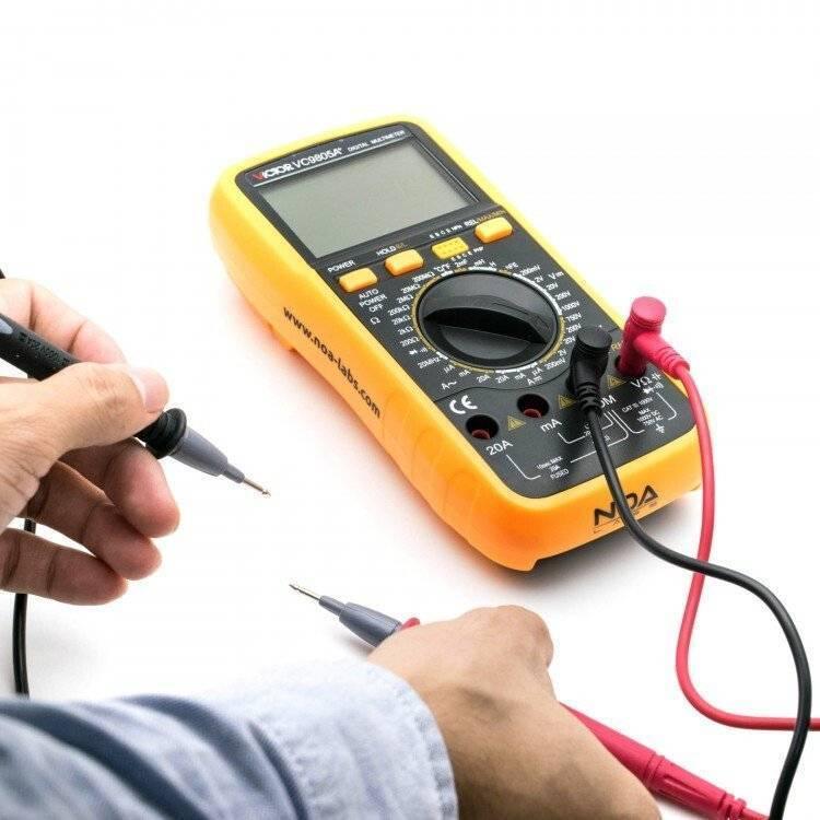 VC9805A+ Digital Multimeter - shown with a blank screen, a central dial, buttons, and red and black test leads held by a person's hands.
