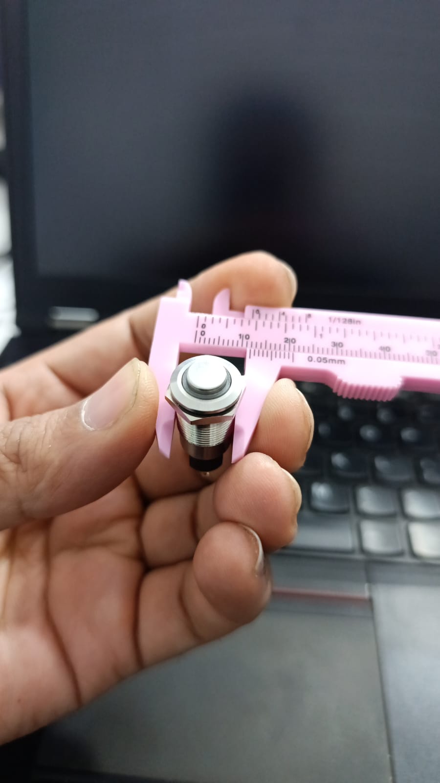 ZLQ-B5V 12mm Waterproof Metal Push ON Push OFF Button Switch With Power Indication - held in a hand and measured by a pink plastic vernier caliper.